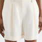 Wearing the TWP Cisco Short—high-waisted, cream pleated shorts in coated viscose linen with belt loops and cuffed hems—this person stands with hands at their sides, showcasing an effortless summer staple.