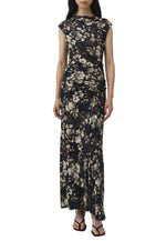 A woman wears the Simkhai Acacia Jersey Midi Dress—a sleeveless, ankle-length dark floral printed dress with a ruched side detail—paired with black sandals, standing against a plain white background.