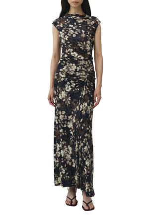 A woman wears the Simkhai Acacia Jersey Midi Dress—a sleeveless, ankle-length dark floral printed dress with a ruched side detail—paired with black sandals, standing against a plain white background.