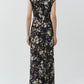 A woman stands facing away, wearing the Simkhai Acacia Jersey Midi Dress—a dark, floral-printed design with short sleeves—paired with heeled sandals against a plain light background.