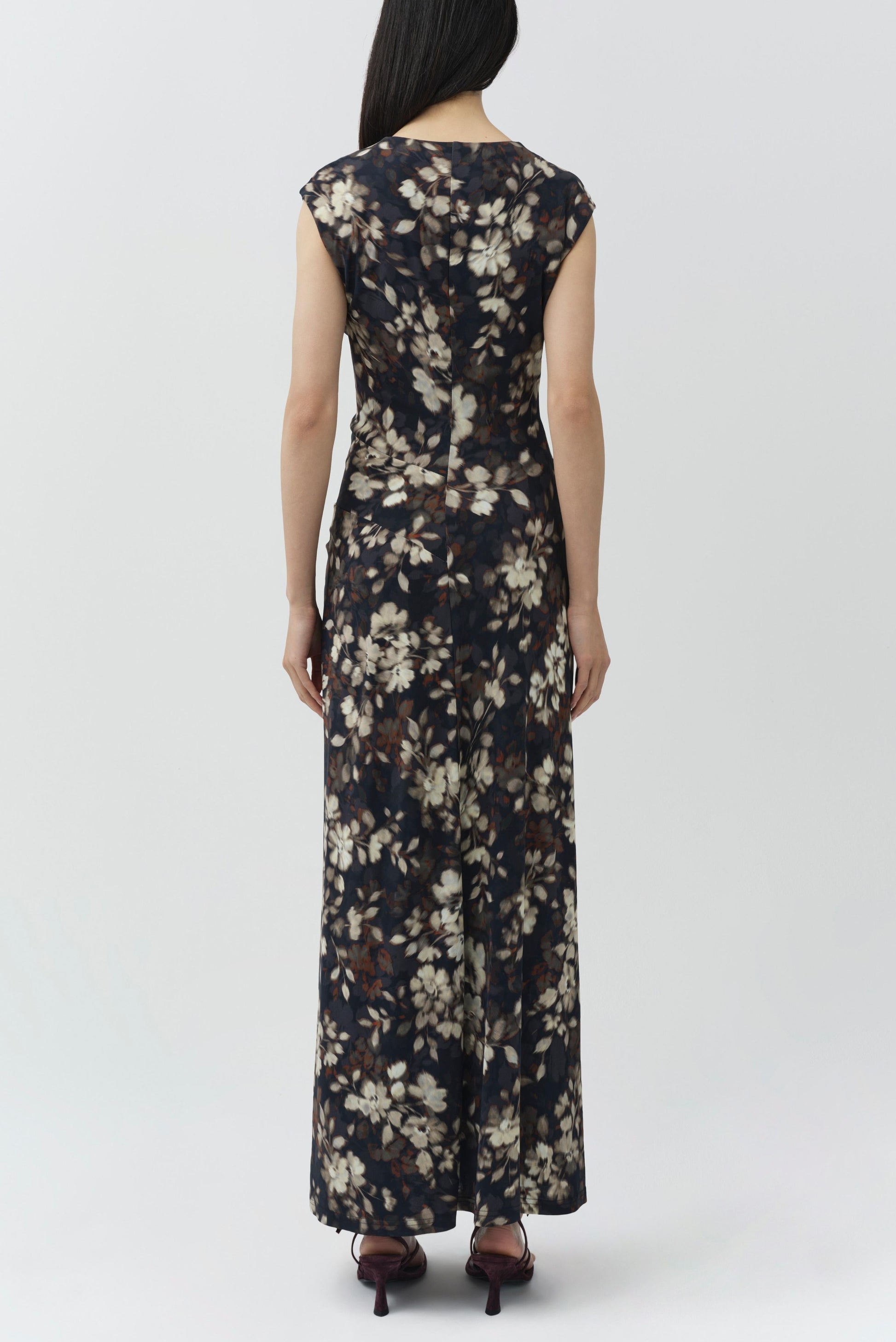 A woman stands facing away, wearing the Simkhai Acacia Jersey Midi Dress—a dark, floral-printed design with short sleeves—paired with heeled sandals against a plain light background.