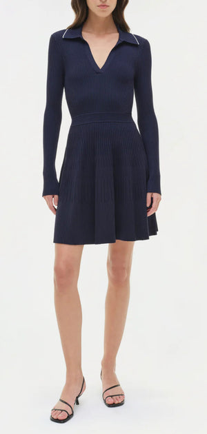 A woman wears the Simkhai Alcott Knit Mini Dress—a navy blue, ribbed knit mini with a spread collar, long sleeves, and flared skirt—paired with black open-toe heeled sandals.