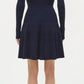 A woman faces away, wearing the Simkhai Alcott Knit Mini Dress in dark navy with long sleeves and pleats, paired with black open-heel sandals, set against a plain white background.