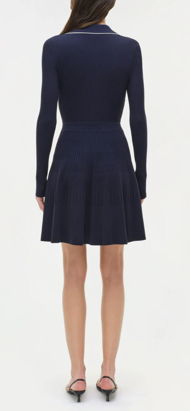 A woman faces away, wearing the Simkhai Alcott Knit Mini Dress in dark navy with long sleeves and pleats, paired with black open-heel sandals, set against a plain white background.