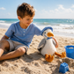 A young boy sits on the sandy beach, smiling at Seymour Seagull—the Mon Ami plush seagull toy wearing a blue hat—while blue bucket and sandcastles sit nearby, with the ocean and sky in the background.