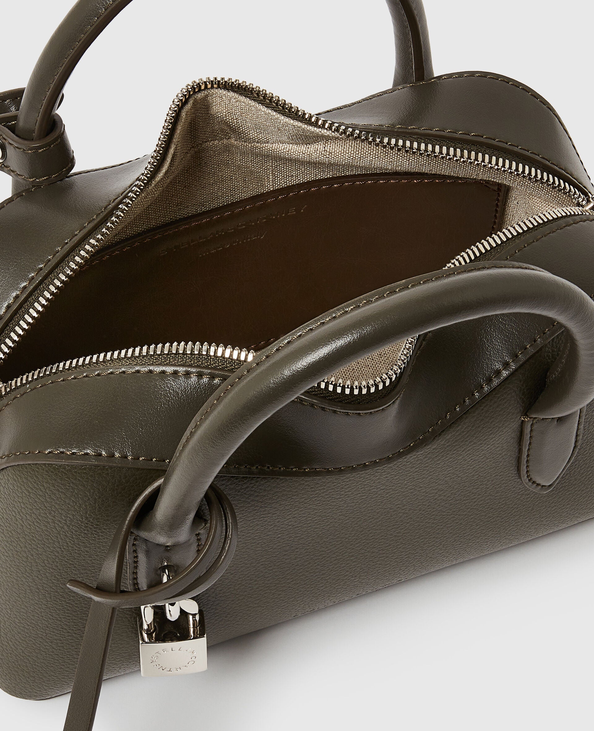 A close-up of the Stella McCartney Ryder Crossbody Bag in olive green, Italian handcrafted with a silver zipper partially open, revealing the fabric interior and a small lock on the handle.