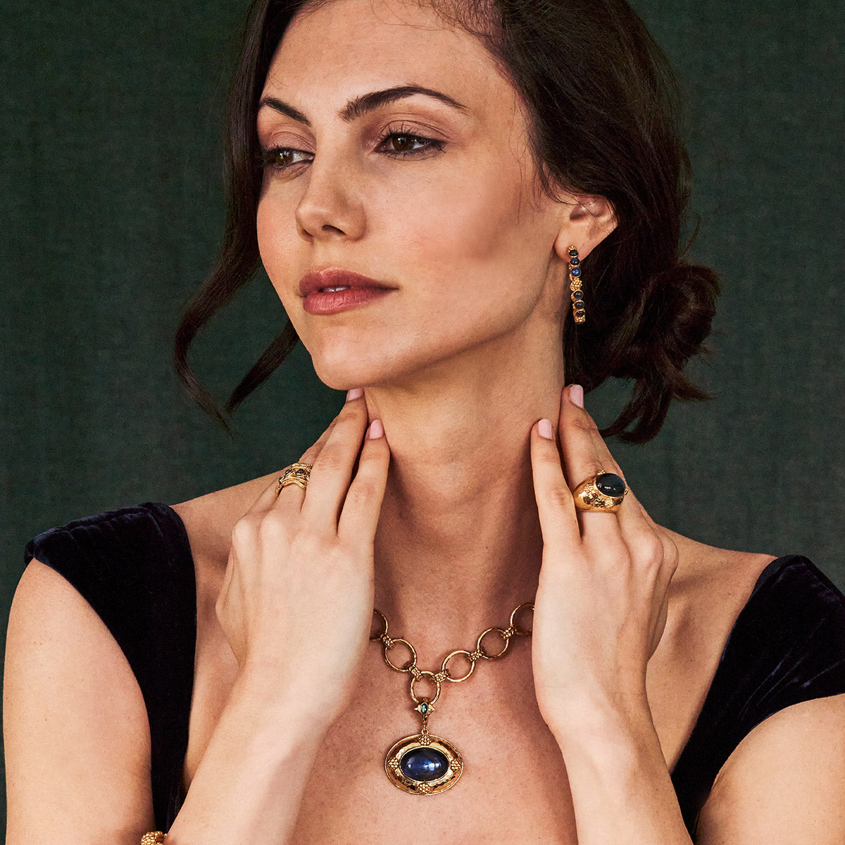 A woman in a black dress poses against a dark background, wearing Juliska Capucine De Wulf's Berry Gem Medium Hoop Earrings along with gold and blue gemstone jewelry—showcasing timeless feminine elegance.