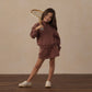 A young girl wears the Rylee & Cru Girls’ Sports Skirt featuring an elastic waistband, posing confidently indoors with a vintage wooden tennis racket resting on her shoulder and standing on a wooden floor for a sporty look.