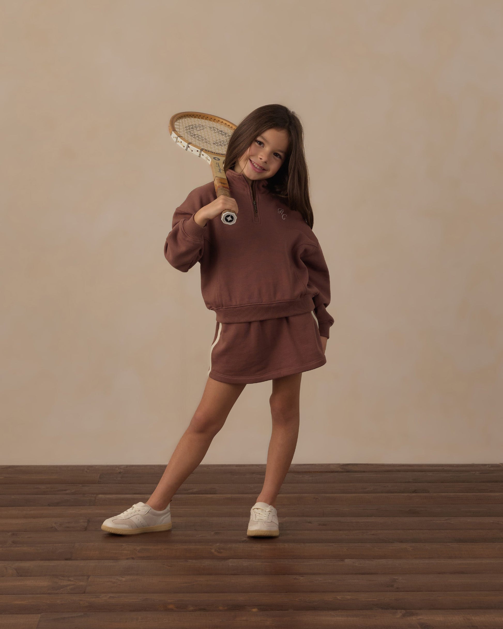 A young girl wears the Rylee & Cru Girls’ Sports Skirt featuring an elastic waistband, posing confidently indoors with a vintage wooden tennis racket resting on her shoulder and standing on a wooden floor for a sporty look.