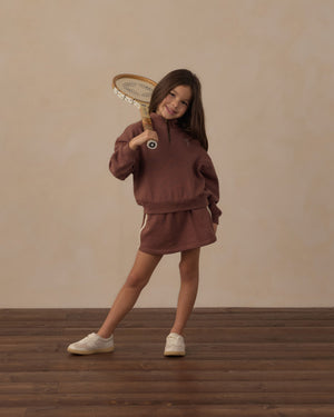 A young girl wears the Rylee & Cru Girls’ Sports Skirt featuring an elastic waistband, posing confidently indoors with a vintage wooden tennis racket resting on her shoulder and standing on a wooden floor for a sporty look.