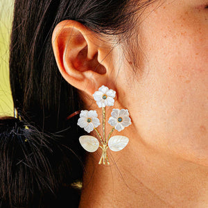 A person wearing Juliska Capucine De Wulf's Mermaid Garden Convertible Bouquet Post Earrings, shaped like white flowers with gold stems and green gemstone centers.