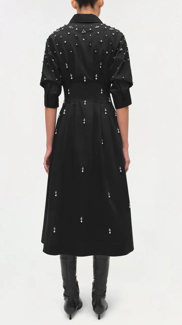 A person stands facing away against a plain background, wearing the Simkhai Jazz Embellished Midi Dress—black cotton poplin with white crystal pearl details—paired with black boots and sleeves rolled up to the elbows.
