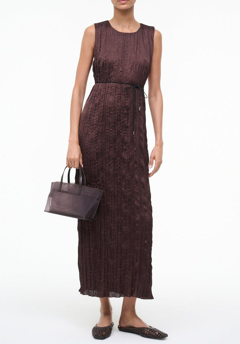 A person in a sleeveless, ankle-length dark brown dress with a thin waist tie holds a small brown handbag and wears Staud Otto Hand-Knotted Flat Mules.