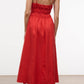 A woman faces away, wearing the Staud Ayla Linen Dress—a sleeveless red midi with thin straps and a gathered back—paired with flat sandals, set against a plain backdrop.