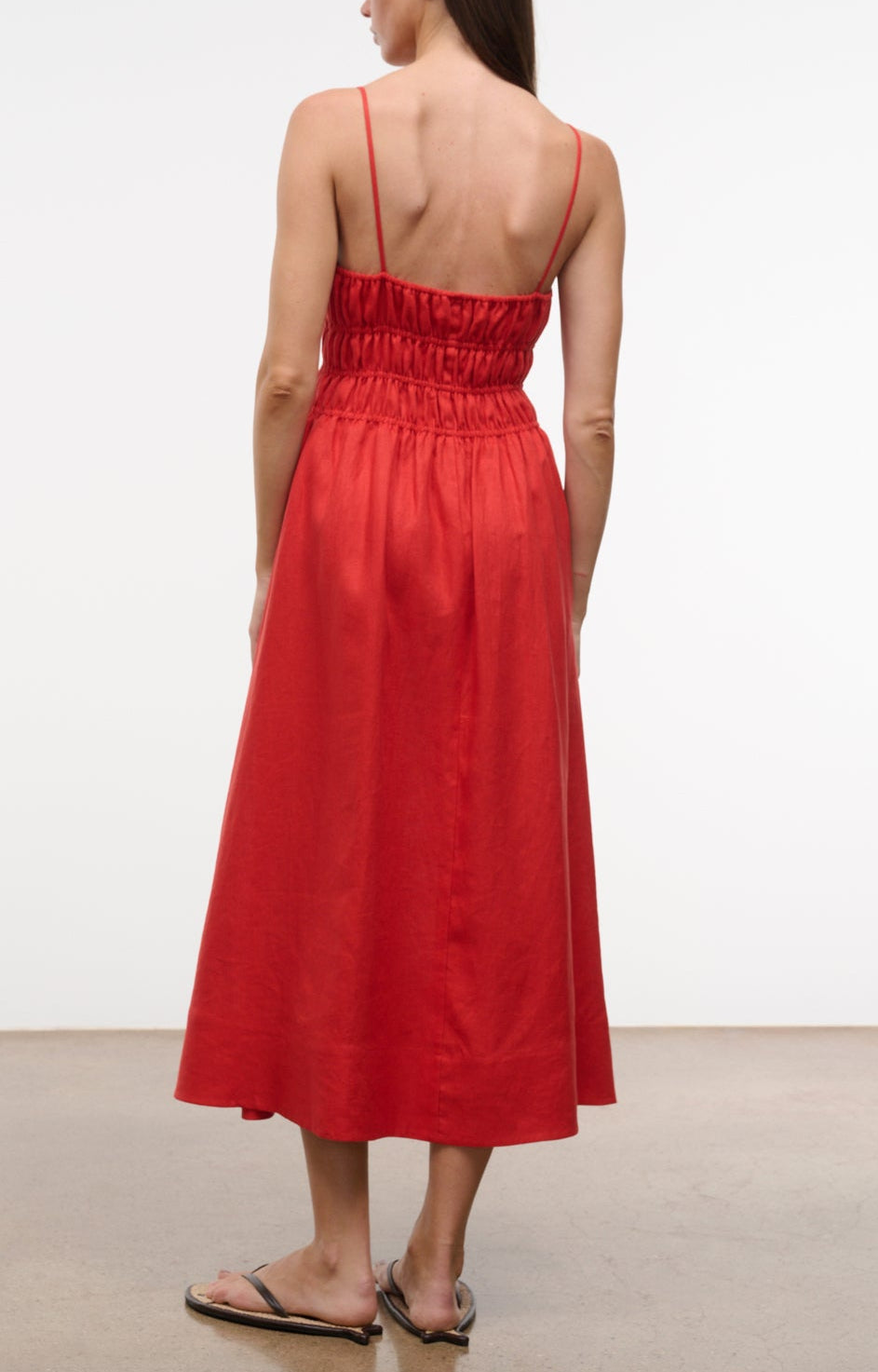 A woman faces away, wearing the Staud Ayla Linen Dress—a sleeveless red midi with thin straps and a gathered back—paired with flat sandals, set against a plain backdrop.