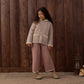 A young girl with long dark hair, dressed in a textured jacket, mauve Rylee & Cru Girls’ Pointelle Knit Pant featuring a ribbed waistband, and slippers stands on a rug in front of a wooden wall with a lantern on the floor beside her.