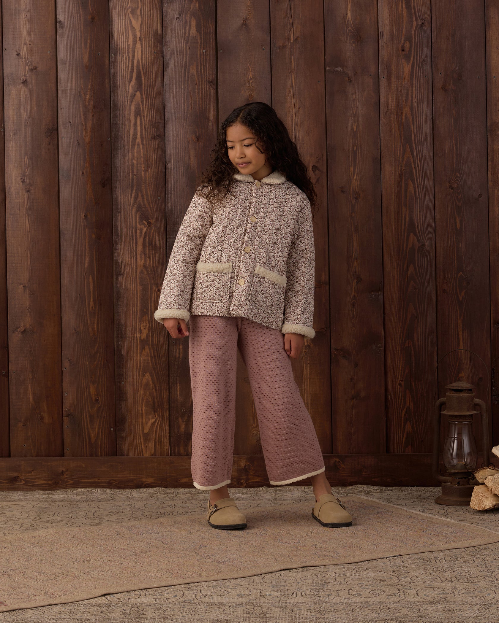 A young girl with long dark hair, dressed in a textured jacket, mauve Rylee & Cru Girls’ Pointelle Knit Pant featuring a ribbed waistband, and slippers stands on a rug in front of a wooden wall with a lantern on the floor beside her.
