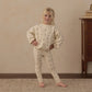 A young child stands indoors on a patterned rug, wearing the Rylee & Cru Baby Girls’ and Girls’ Spongey Knit Set—a cream-colored ensemble with small prints featuring snug leggings and a cozy pullover sweatshirt—beside a wooden dresser topped with stacked books.