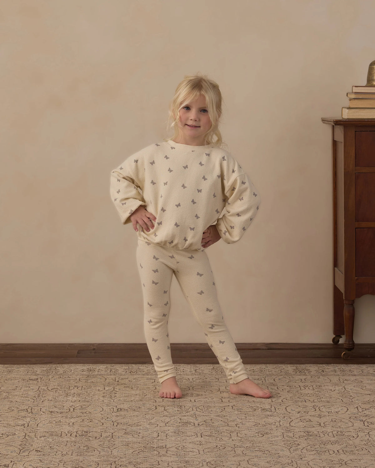 A young child stands indoors on a patterned rug, wearing the Rylee & Cru Baby Girls’ and Girls’ Spongey Knit Set—a cream-colored ensemble with small prints featuring snug leggings and a cozy pullover sweatshirt—beside a wooden dresser topped with stacked books.
