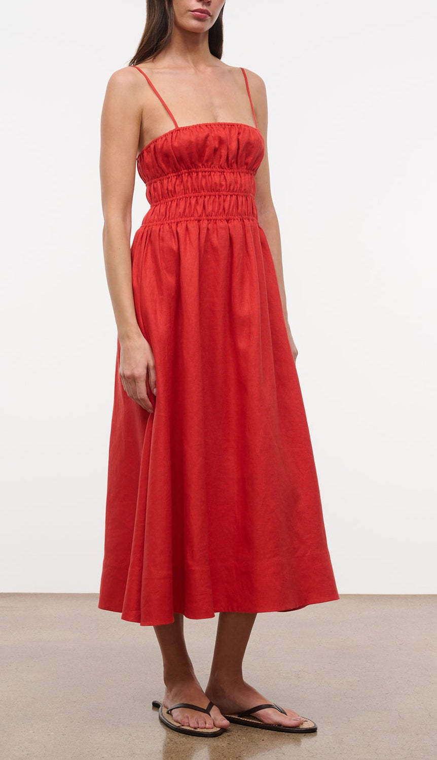 A woman stands on a plain floor in flip-flops, wearing the Staud Ayla Linen Dress—a sleeveless, ankle-length red dress with thin straps and a gathered waist.