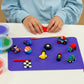 A child in a blue shirt uses the Floss and Rock Cars Clay & Play set, molding red air-dry clay at a table with small colorful clay vehicles, sculpting tools, and tubs of modeling compound on a purple mat.