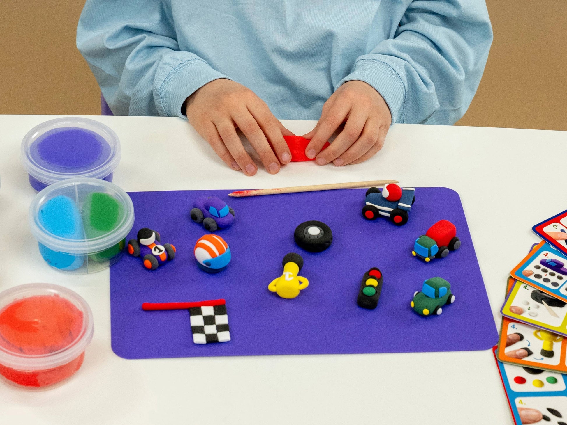 A child in a blue shirt uses the Floss and Rock Cars Clay & Play set, molding red air-dry clay at a table with small colorful clay vehicles, sculpting tools, and tubs of modeling compound on a purple mat.