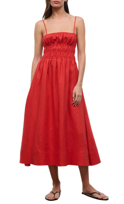 A woman stands against a white background wearing the Staud Ayla Linen Dress—a sleeveless red midi with thin straps and gathered bodice—paired with black flip-flops.
