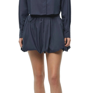 Model wearing the Simkhai Pippa Bubble Mini Skirt in navy blue, styled with a matching long-sleeve cotton poplin top for a voluminous look, photographed from shoulders to mid-thigh against a white background.