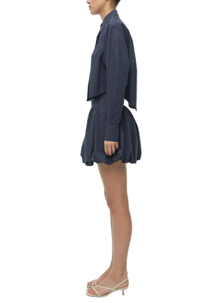 A woman stands in profile against a white background, wearing Simkhai's navy blue cotton poplin Pippa Bubble Mini Skirt, a matching long-sleeve shirt, and white open-toe heels.