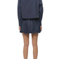 A person wearing the Simkhai Pippa Bubble Mini Skirt in navy stands facing away, paired with a matching long-sleeve jacket, clear-heeled shoes, and bare legs against a white background.