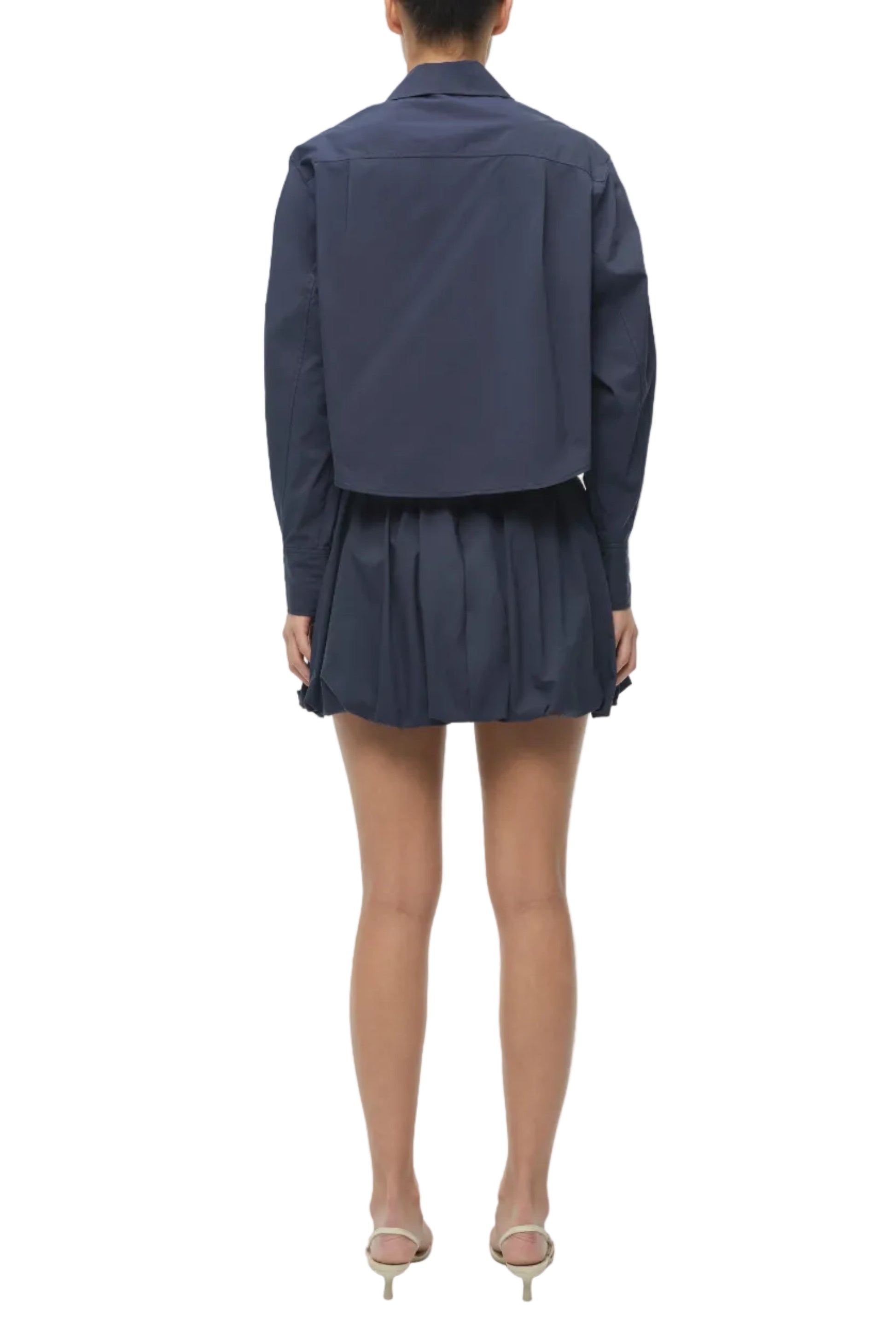 A person wearing the Simkhai Pippa Bubble Mini Skirt in navy stands facing away, paired with a matching long-sleeve jacket, clear-heeled shoes, and bare legs against a white background.