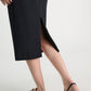 A person wears the Dorothee Schumacher Emotional Essence Skirt—black Punto Milano with a back slit—paired with black flat sandals on a light floor. Only the lower body is visible, showcasing this effortless workweek essential.