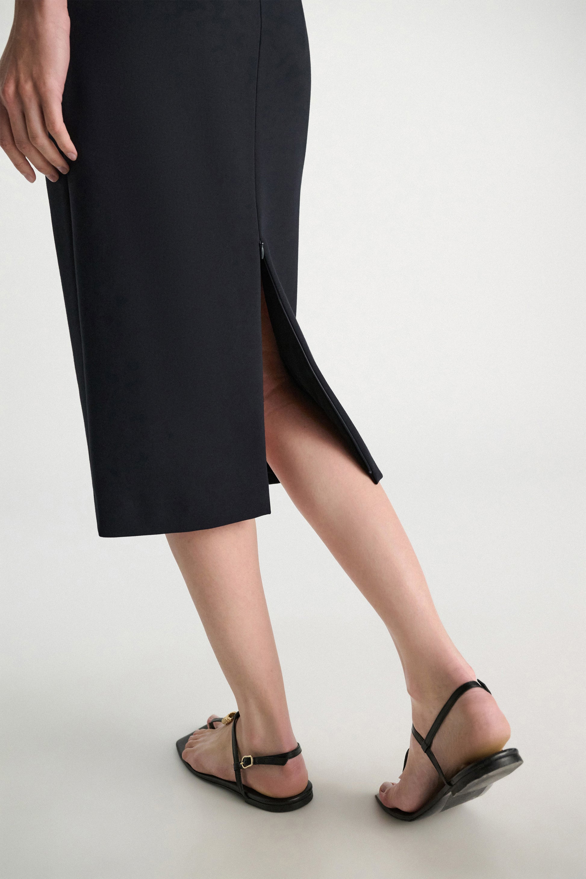 A person wears the Dorothee Schumacher Emotional Essence Skirt—black Punto Milano with a back slit—paired with black flat sandals on a light floor. Only the lower body is visible, showcasing this effortless workweek essential.