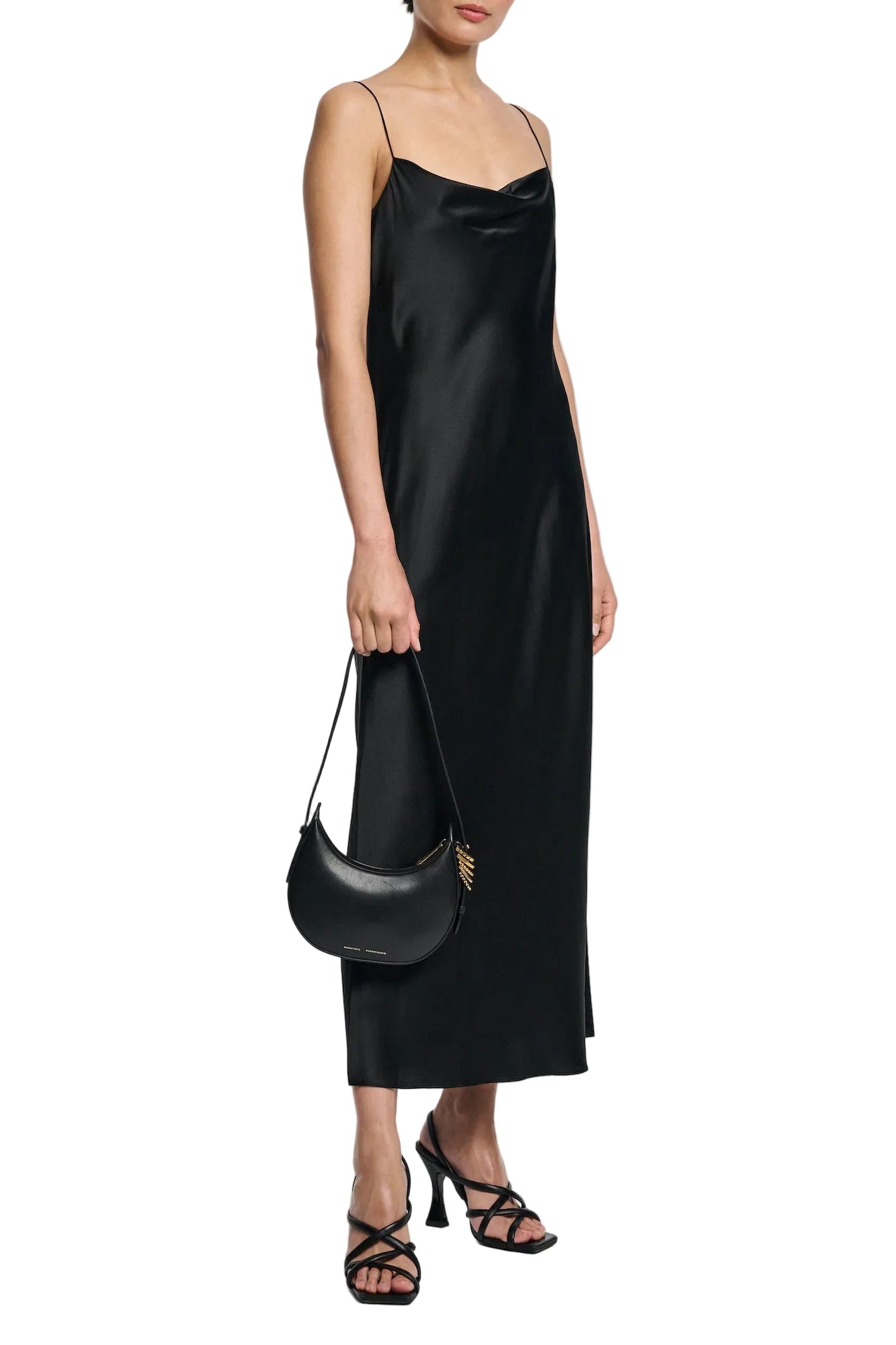 A woman wears the Sense of Shine Dress by Dorothee Schumacher, a stretch silk-satin midi with adjustable spaghetti straps, paired with black strappy heeled sandals and a small black handbag.