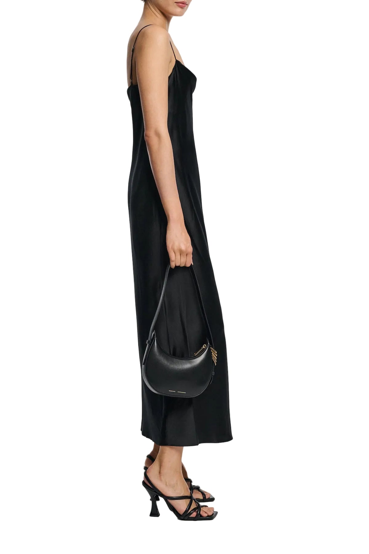 A woman in the Dorothee Schumacher Sense of Shine Dress, a stretch silk-satin midi with adjustable spaghetti straps, stands in profile wearing black strappy heels and holding a small black shoulder bag.