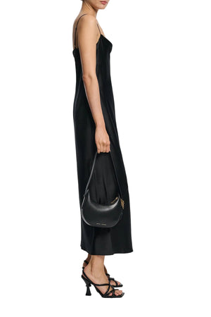 A woman in the Dorothee Schumacher Sense of Shine Dress, a stretch silk-satin midi with adjustable spaghetti straps, stands in profile wearing black strappy heels and holding a small black shoulder bag.