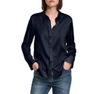 A person wears the Dorothee Schumacher Sense of Shine Blouse, a long-sleeve dark navy stretch silk satin shirt with a classic collar, paired with blue jeans, standing hands in pockets against a white background.