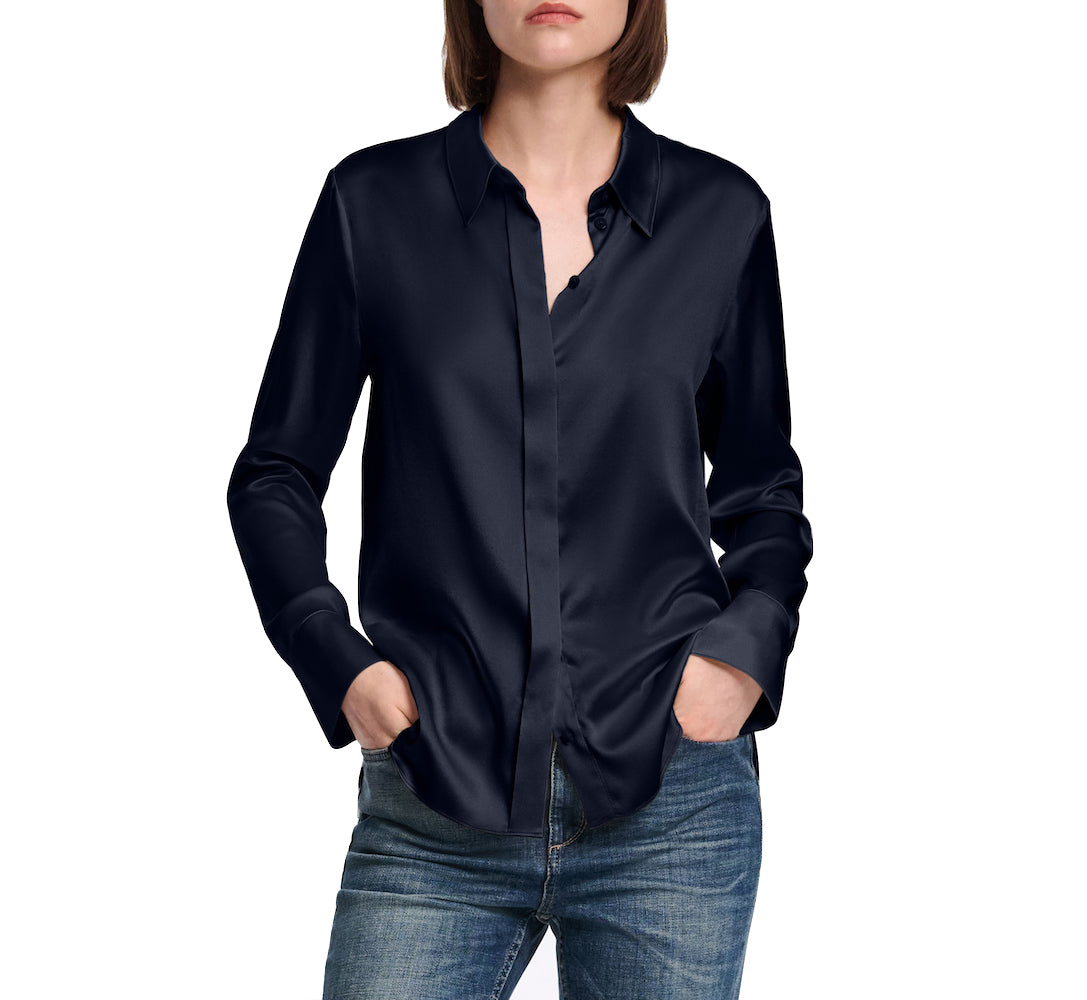 A person wears the Dorothee Schumacher Sense of Shine Blouse, a long-sleeve dark navy stretch silk satin shirt with a classic collar, paired with blue jeans, standing hands in pockets against a white background.