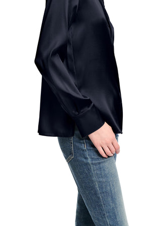 A person wears the Dorothee Schumacher Sense of Shine Blouse in fitted black stretch silk satin with a classic collar and blue jeans, standing with their left hand by their side against a white background.