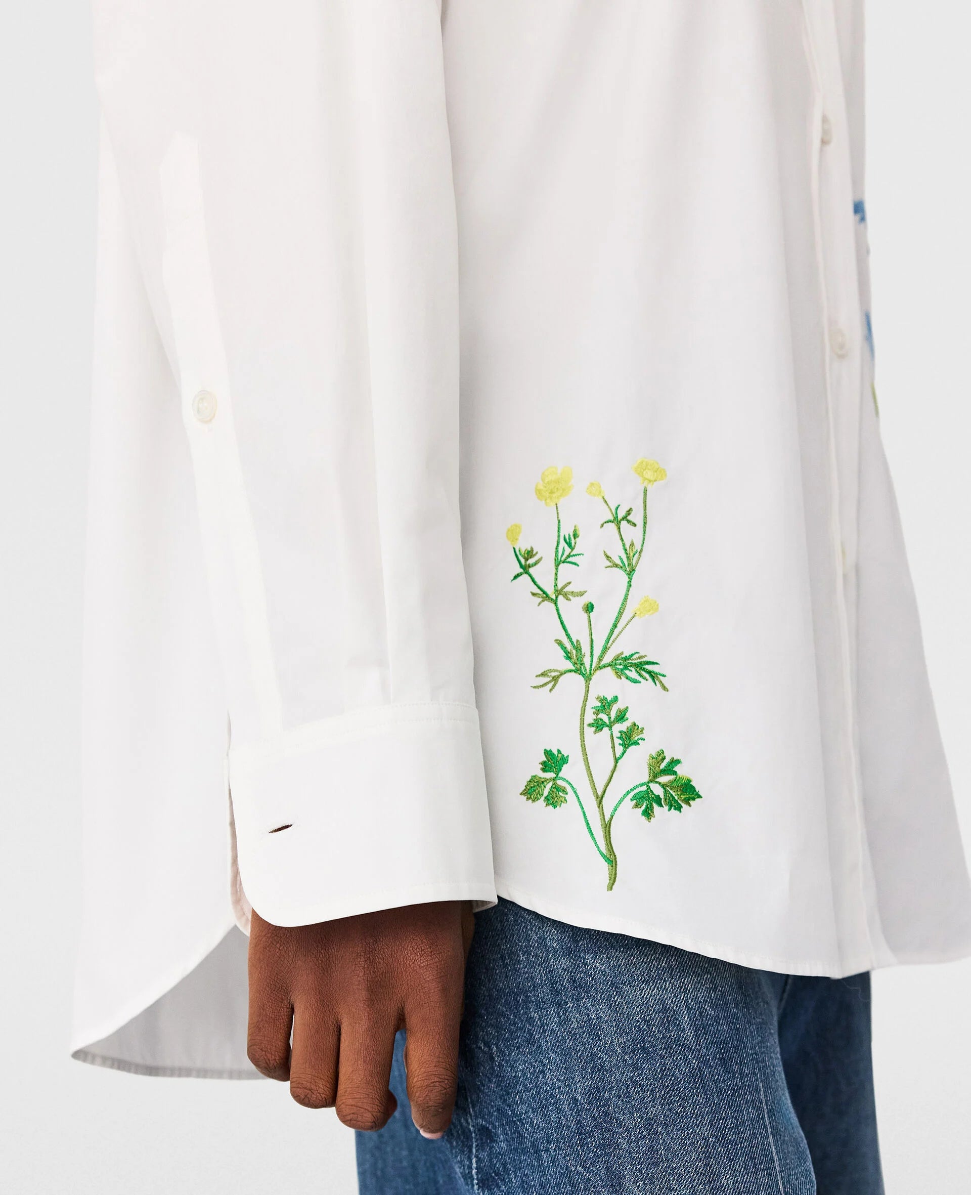A person wearing the Stella McCartney Flower Embroidered Shirt with yellow floral designs near the hem, paired with blue jeans.