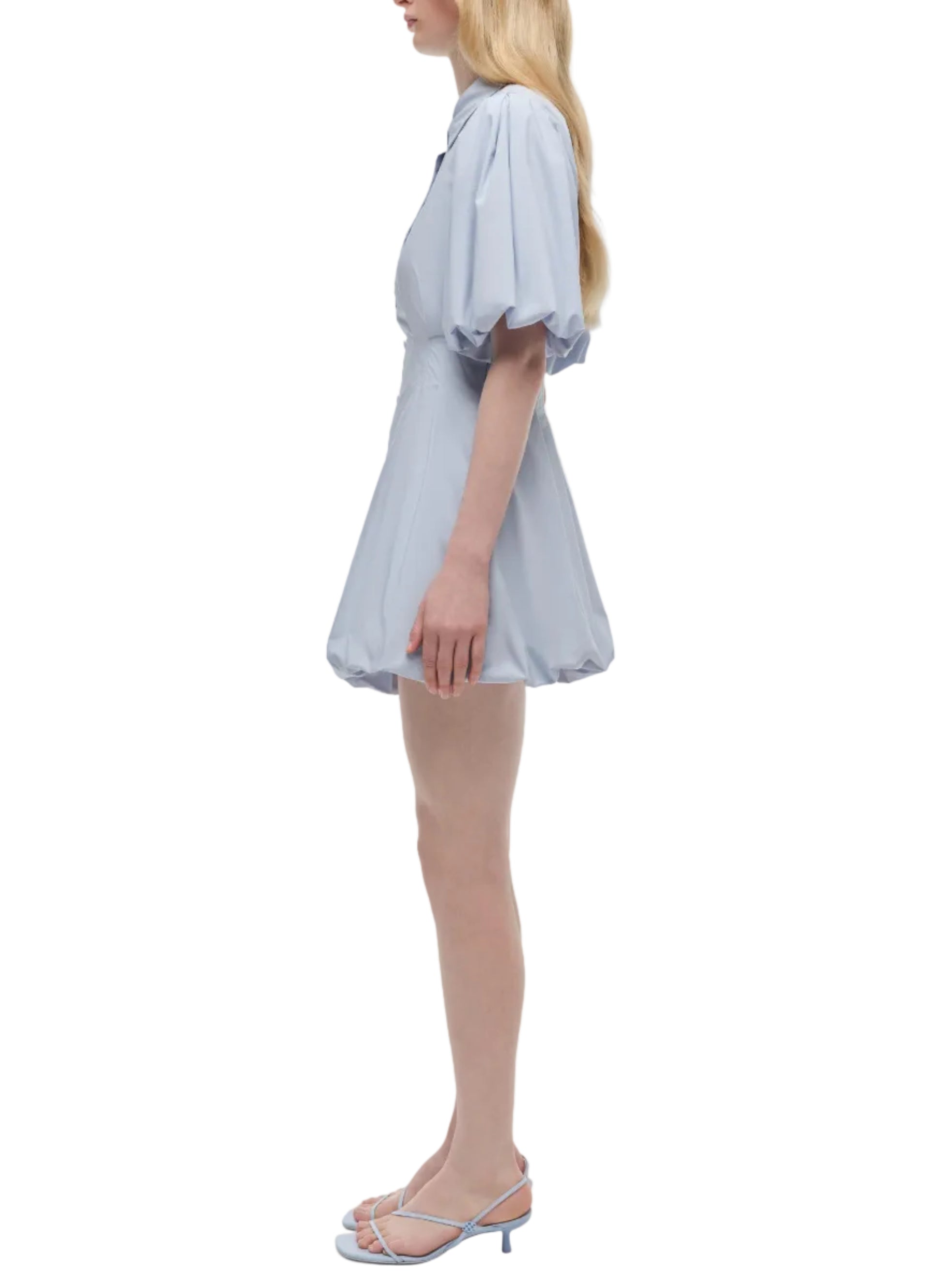 Woman in profile wears the Simkhai Cleo Dress, a light blue cotton mini dress with balloon sleeves, paired with matching strappy heeled sandals against a white background.