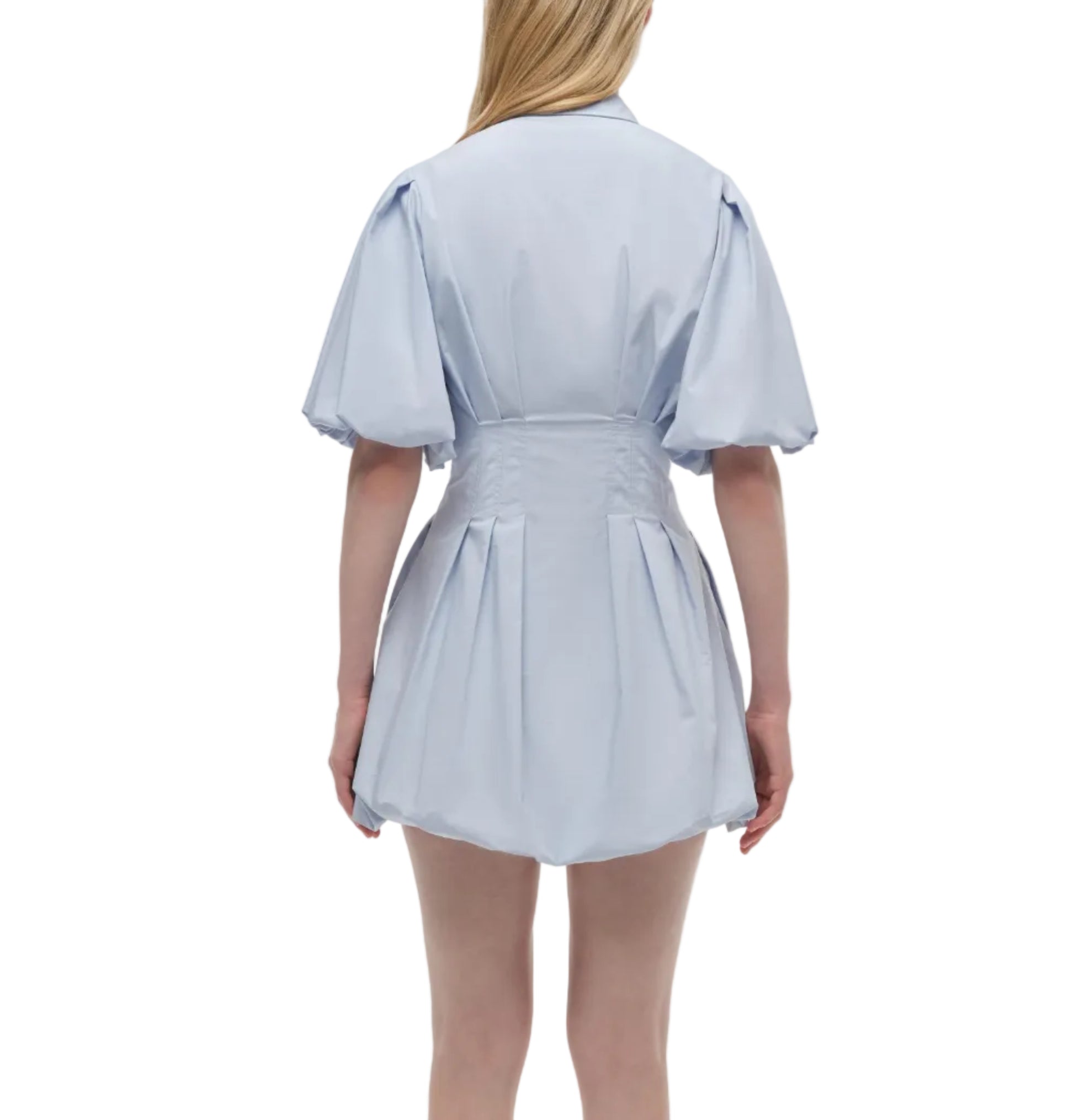 A woman faces away in the Simkhai Cleo Dress, a light blue cotton mini with pleats and a belted waist, set against a white background.