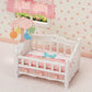 The Calico Critters Crib with Mobile, featuring a small white and pink design with a mobile and pink blanket, sits in a room adorned with floral wallpaper and a window dressed with a checkered curtain, making it a delightful addition to any dollhouse furniture collection or baby nursery set.
