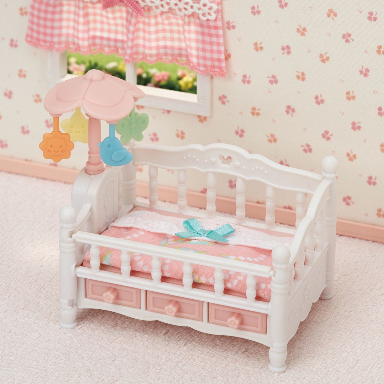 The Calico Critters Crib with Mobile, featuring a small white and pink design with a mobile and pink blanket, sits in a room adorned with floral wallpaper and a window dressed with a checkered curtain, making it a delightful addition to any dollhouse furniture collection or baby nursery set.