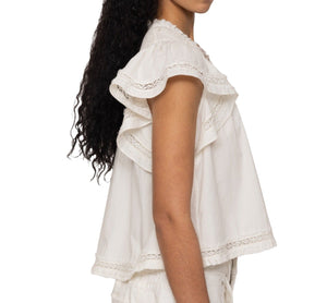 A woman with long curly hair models the Sea NY Lilith Top by Sea NY, a white, cotton, short-sleeved blouse featuring flutter sleeves and ruffle detailing with lace trim, viewed from the side.