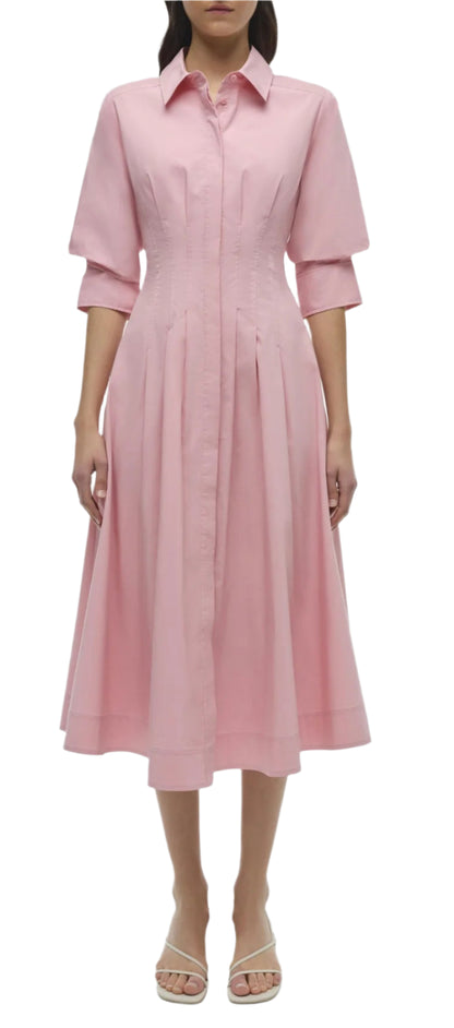 A person wearing the Simkhai Signature Jazz Dress—a pink, button-up cotton poplin dress featuring pintuck midi design and three-quarter sleeves—paired with beige sandals.