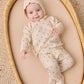 A newborn baby in the Quincy Mae Baby Girls’ Take Home Set, made from cream-colored waffle knit fabric with delicate patterns, lies on a beige mattress in a woven basket, gazing up with a gentle smile—an ideal choice for a cozy first outfit or a newborn coming home ensemble.