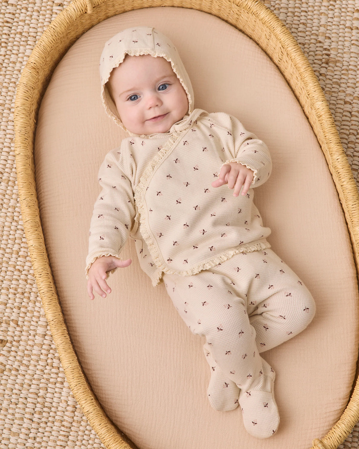 A newborn baby in the Quincy Mae Baby Girls’ Take Home Set, made from cream-colored waffle knit fabric with delicate patterns, lies on a beige mattress in a woven basket, gazing up with a gentle smile—an ideal choice for a cozy first outfit or a newborn coming home ensemble.