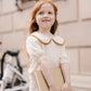 A young girl with long red hair, dressed in a cream-colored Rylee & Cru Girls' Pointelle Collared Top, stands outdoors by a bicycle holding a closed book and smiling slightly in her vintage-inspired outfit.
