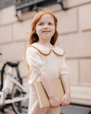 A young girl with long red hair, dressed in a cream-colored Rylee & Cru Girls' Pointelle Collared Top, stands outdoors by a bicycle holding a closed book and smiling slightly in her vintage-inspired outfit.